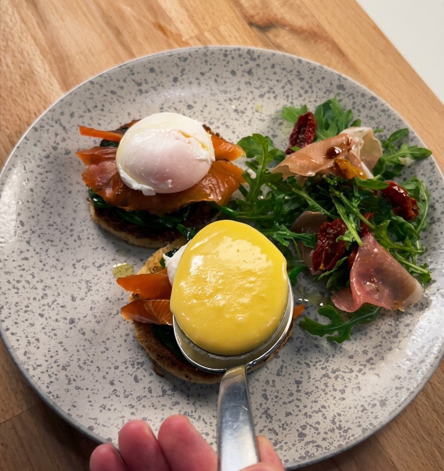 Nahaufnahme von einem großen Löffel, der dicke, glänzende Sauce Hollandaise über ein pochiertes Ei gießt, das auf Räucherlachs und Brioche liegt.