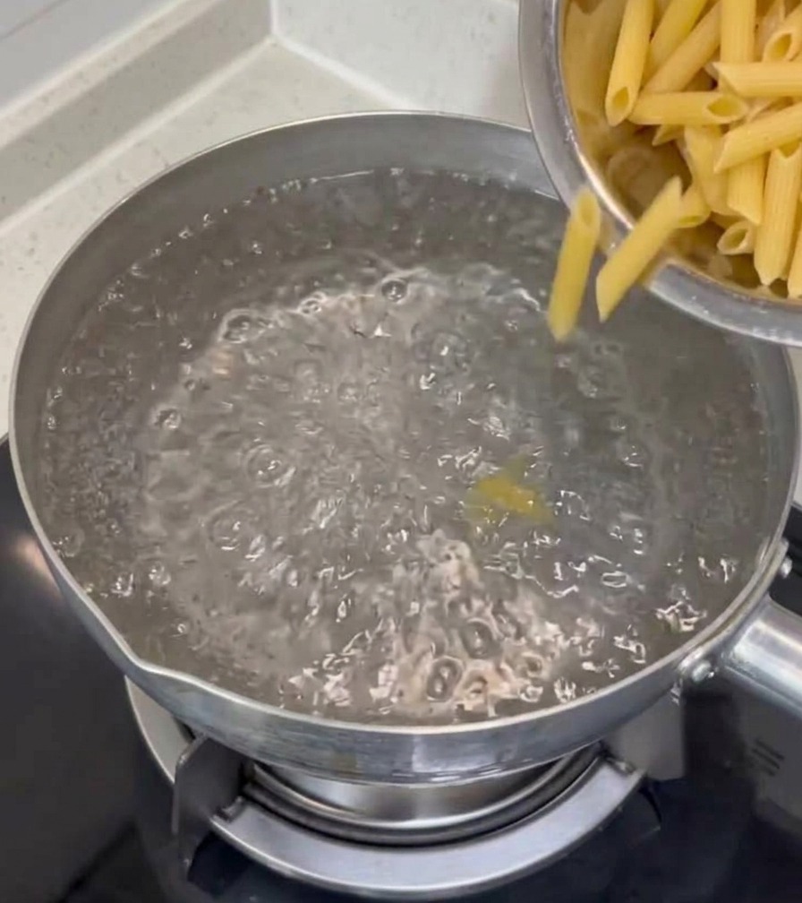 Trockene Penne-Nudeln werden aus einer Edelstahlschüssel in einen Topf mit sprudelnd kochendem Wasser auf dem Herd geschüttet.