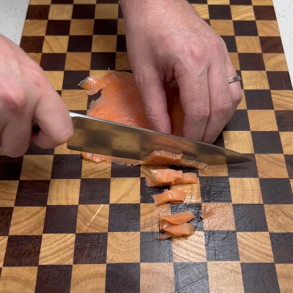 Die Hände eines Kochs schneiden mit einem silbernen Messer flache Stücke Räucherlachs in kleine Quadrate auf einem braun-beige karierten Schneidebrett.