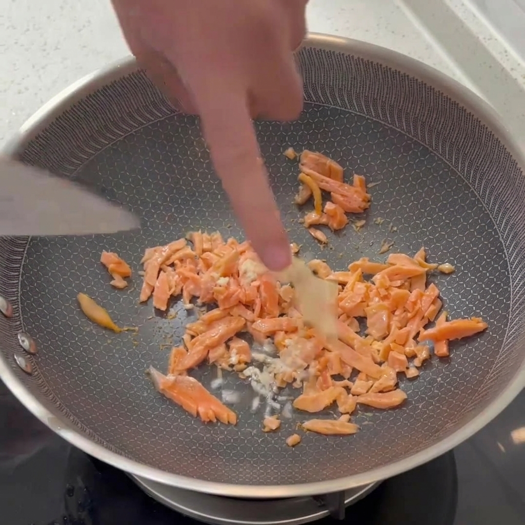 Fein gehackter weißer Knoblauch wird mit einem Pfannenwender unter die angebratenen Räucherlachsfeile gemischt.