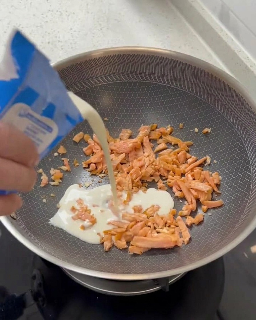 Eine Hand gießt weiße Sahne aus einem blau-weißen Karton in eine Pfanne mit gebratenen Lachsstrücken.