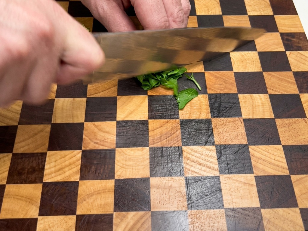 Nahaufnahme von Händen, die mit einem Kochmesser frische grüne Petersilienblätter auf einem karierten Holzschneidebrett fein hacken.