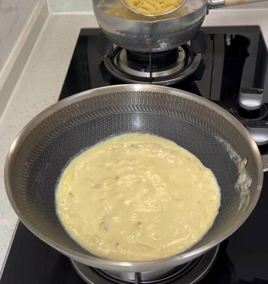 Ein Schaumlöffel aus Metall hebt gekochte Penne-Nudeln aus einem kochenden Topf direkt in eine Pfanne mit cremiger weißer Sauce auf dem Herd.