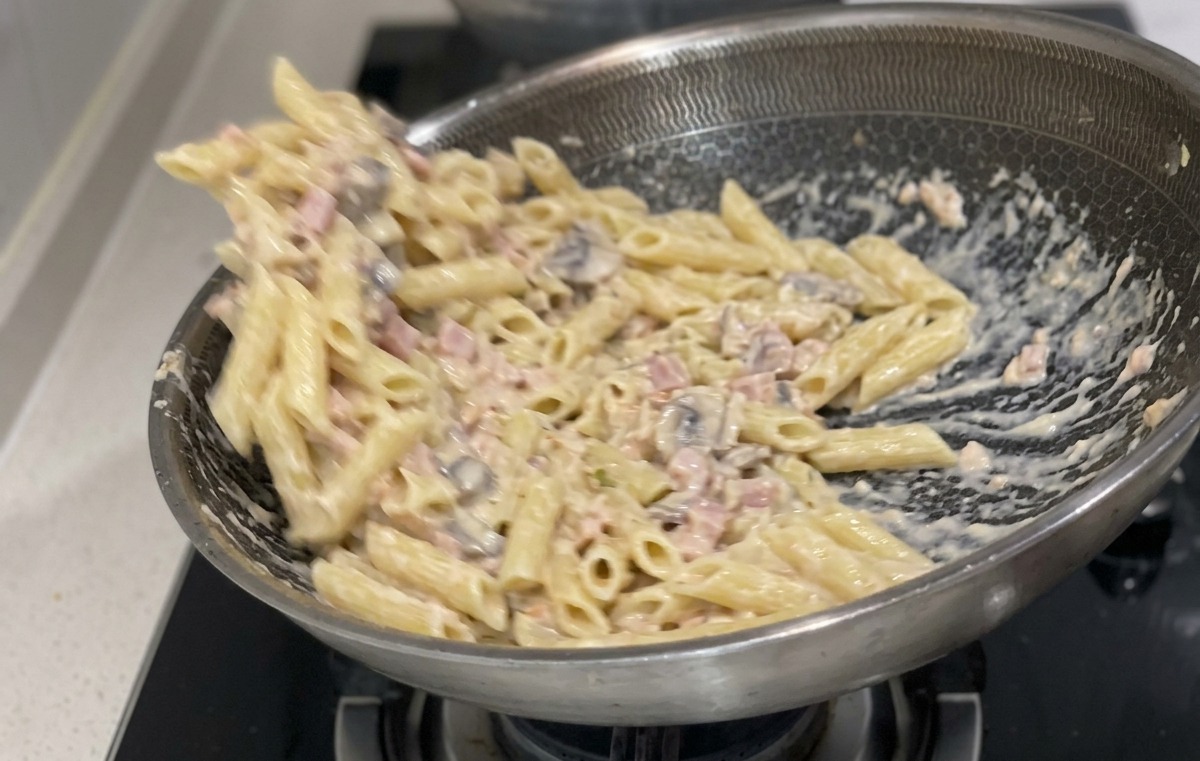 Gekochte Penne-Nudeln werden in einer großen Edelstahlpfanne mit einer cremigen weißen Sauce und Lachsstreifen vermengt.