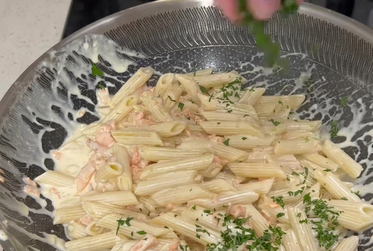 Eine Hand streut fein gehackte grüne Petersilie über eine große Pfanne mit Penne-Nudeln in cremiger weißer Sauce mit rosa Räucherlachsstreifen.