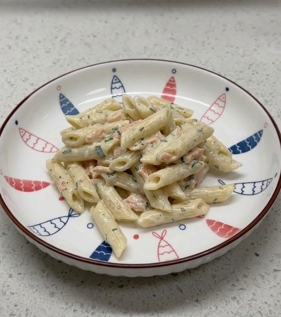 Eine Portion cremige Lachs-Penne auf einem weißen Keramikteller, der am Rand mit einem blau-roten Fischmuster verziert ist.