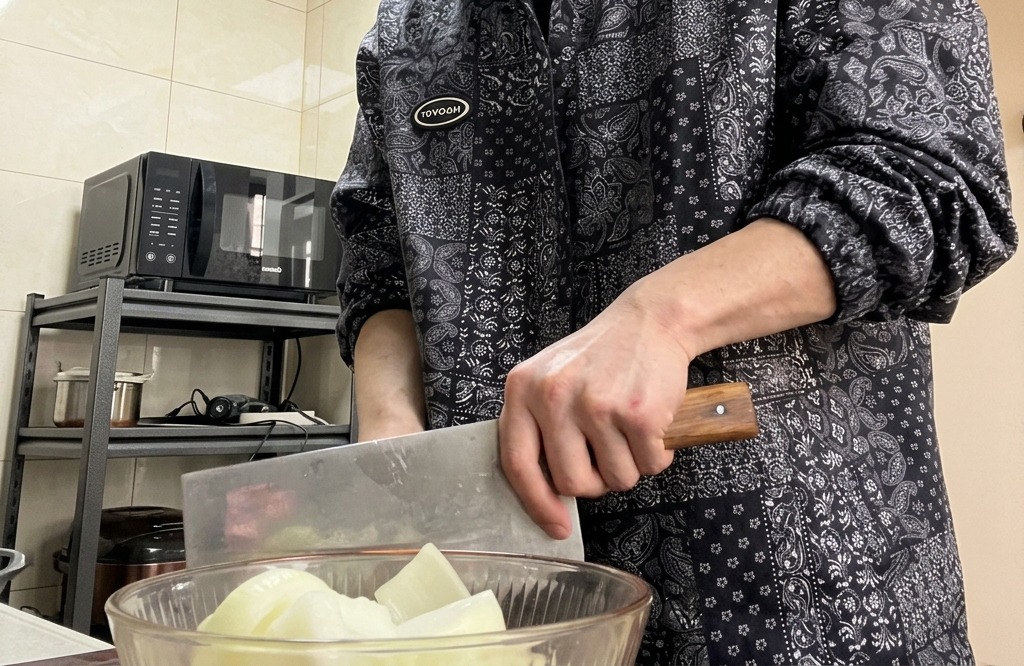 Hände halten ein großes Hackmesser und schneiden eine Zwiebel auf einem Holzschneidebrett.