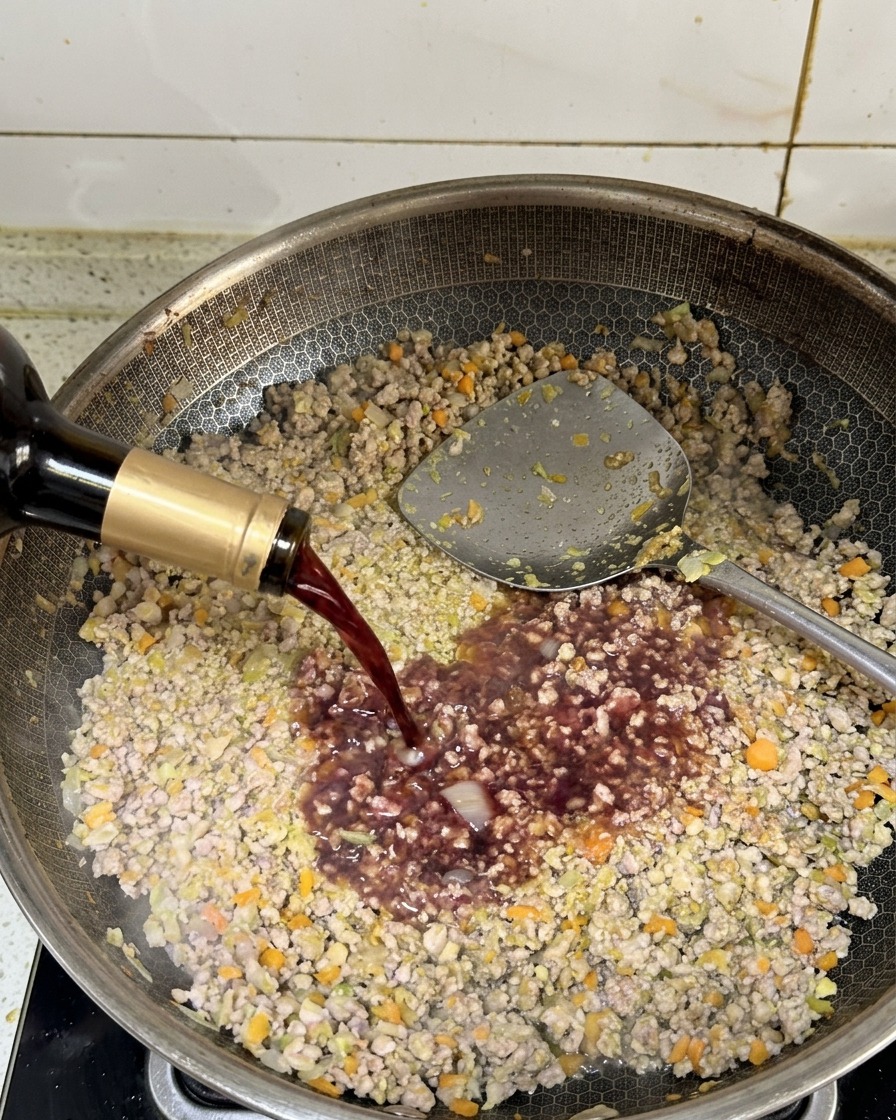 Dunkler Rotwein wird aus einer Flasche in eine Pfanne mit angebratenem Hackfleisch gegossen.