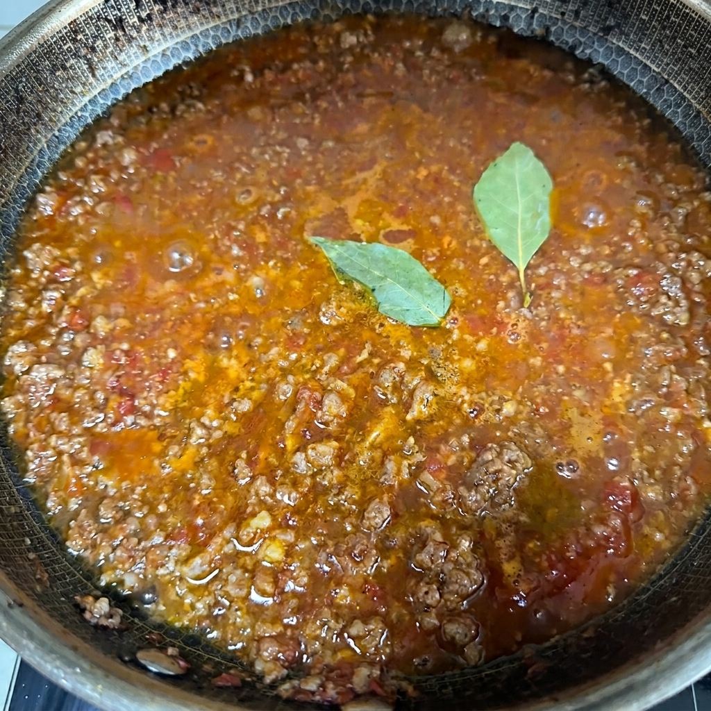 Nahaufnahme einer blubbernden, reichhaltigen Fleischsauce, die in einer großen Pfanne köchelt, während zwei frische grüne Lorbeerblätter auf der Oberfläche liegen.
