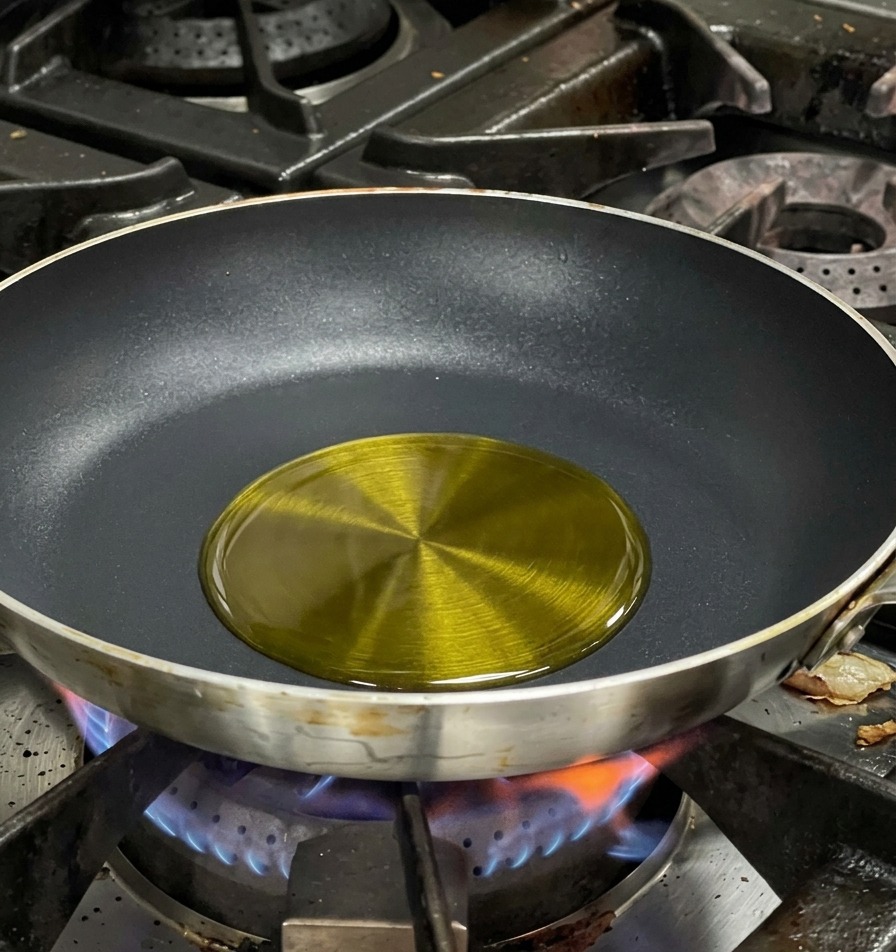 Schimmerndes Olivenöl in der Mitte einer schwarzen beschichteten Bratpfanne über einer blauen Gasflamme.