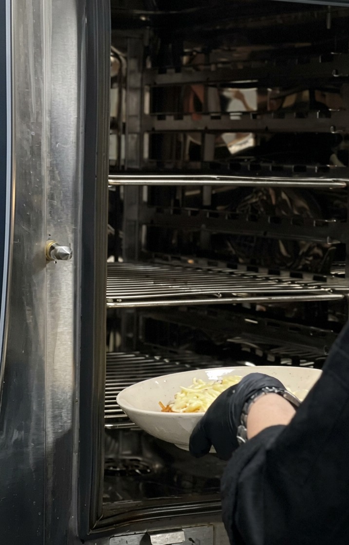 Ein Koch mit schwarzen Handschuhen schiebt eine weiße, mit Käse bestreute Schale Pasta in einen großen industriellen Edelstahlofen.