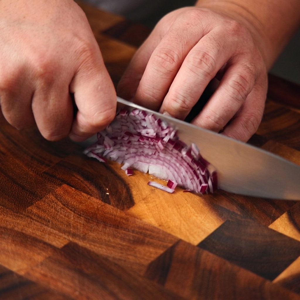 Nahaufnahme von Händen, die eine lila Zwiebel mit einem Kochmesser auf einem Holzbrett fein würfeln.