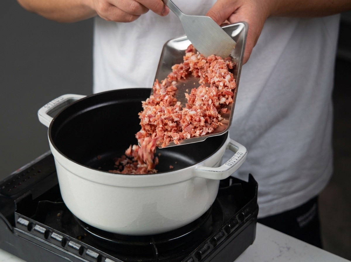 Gewürfelte Pancetta wird von einem Metalltablett in einen weißen, emaillierten gusseisernen Topf auf einem tragbaren Herd geschüttet.