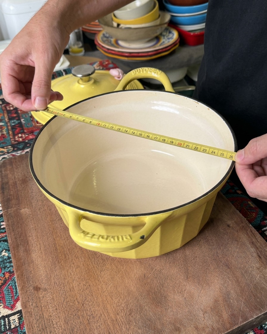 Eine Person misst den oberen Durchmesser eines gelben Bräters mit einem gelben Maßband, um zu bestätigen, dass er etwa 26 Zentimeter breit ist.