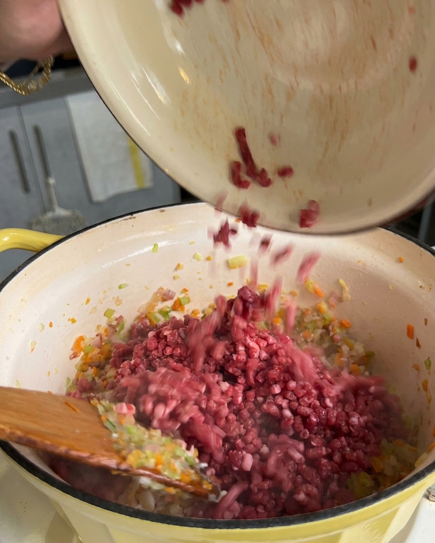 Hackfleisch wird aus einer Schüssel in einen Topf mit angeschwitzten Karotten-, Sellerie- und Zwiebelwürfeln gegeben.