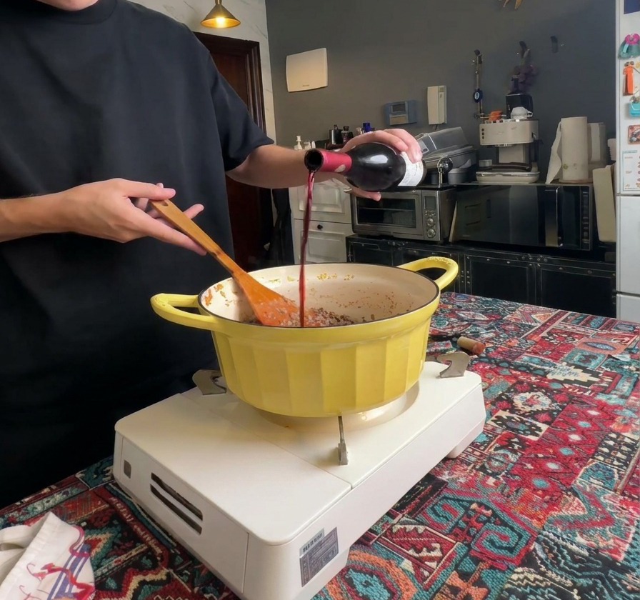 Rotwein wird aus einer dunklen Glasflasche in einen Topf mit gut gebräuntem Hackfleisch gegossen.