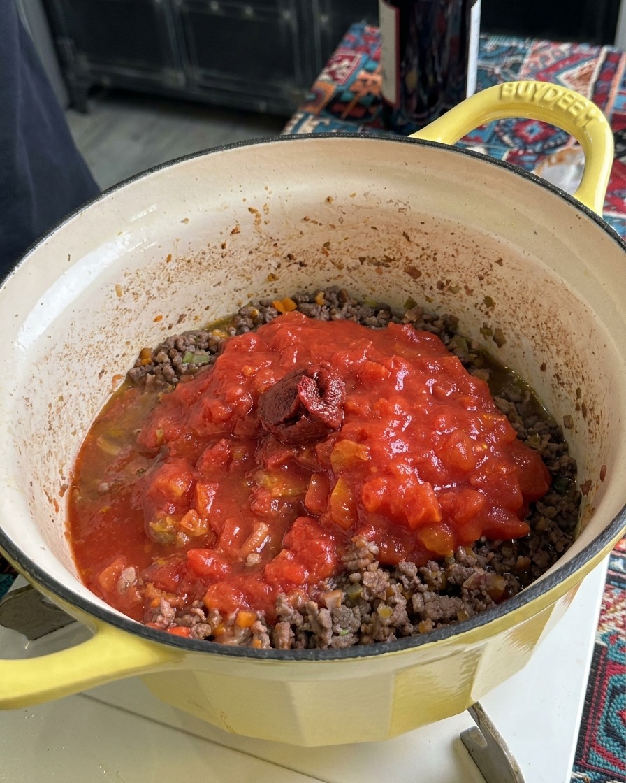 Ein Klecks dickflüssiges, dunkles Tomatenmark liegt auf passierten Tomaten und Hackfleisch in einem gelben Topf.