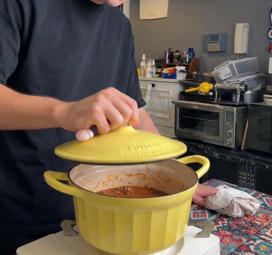 Eine Person setzt einen gelben Gusseisendeckel auf einen Topf mit Ragù, um den langsamen Schmorprozess zu starten.
