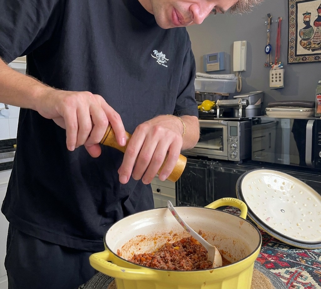 Eine Person verwendet eine hölzerne Pfeffermühle, um frischen schwarzen Pfeffer in eine dicke italienische Fleischsauce in einem gelben Bräter zu mahlen.