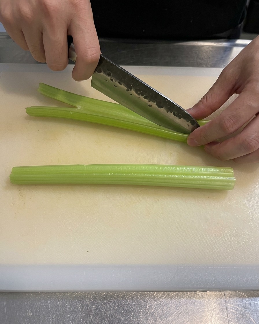 Nahaufnahme einer Hand, die mit einem scharfen Messer grüne Staudenselleriestangen auf einem weißen Schneidebrett in kleine Stücke schneidet.