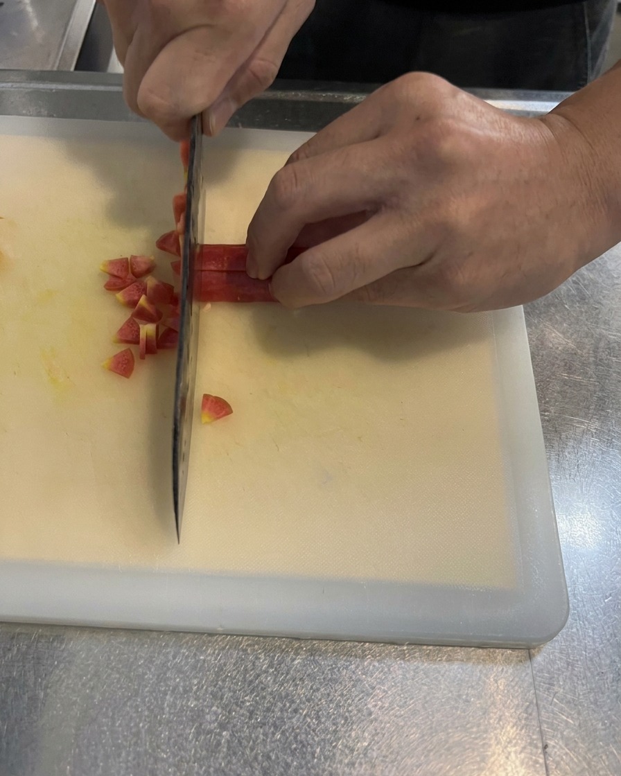 Hände würfeln eine leuchtende Karotte mit einem Küchenmesser in kleine, gleichmäßige Quadrate auf einem weißen Schneidebrett.