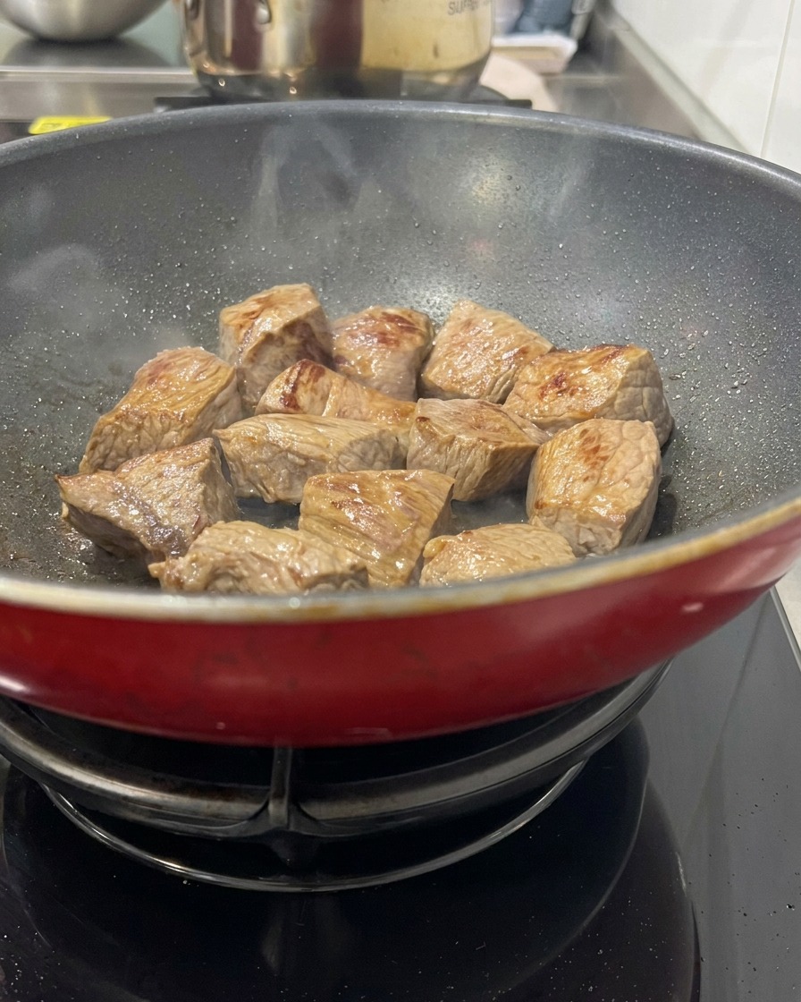 Gewürfeltes Rindfleisch brät in einer heißen roten Pfanne an, zeigt goldbraun versiegelte Oberflächen und leichter Dampf steigt auf.
