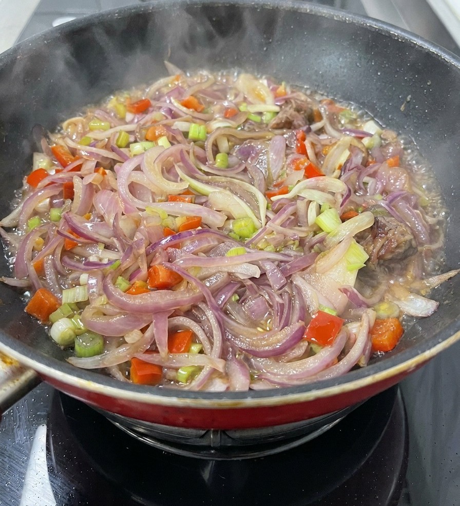 Eine Pfanne voller geschnittener roter Zwiebeln, Karotten, Sellerie und Fleischstücken köchelt in etwas Flüssigkeit, während Dampf aufsteigt.