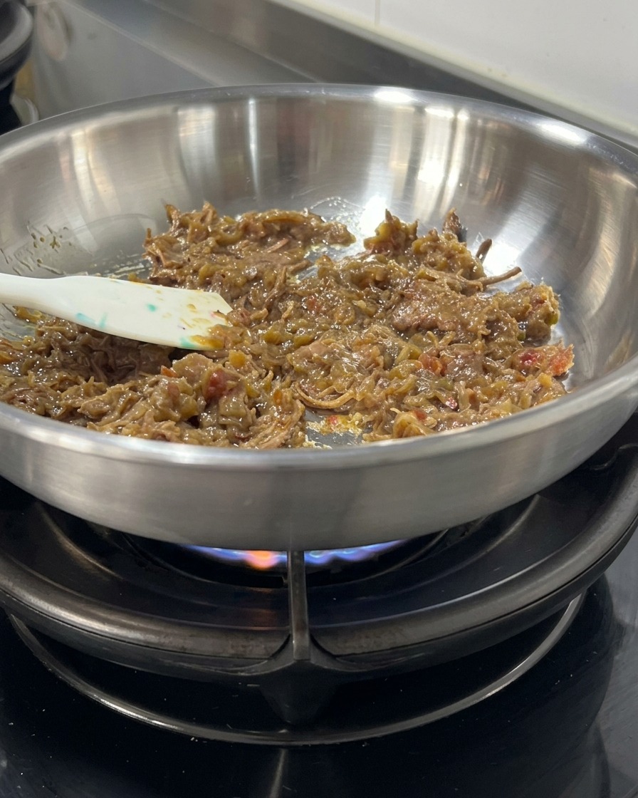 Dicke, dunkle Fleischsauce und zerzupftes Rindfleisch werden mit einem Spatel in einer Edelstahlpfanne über einer Flamme umgerührt.