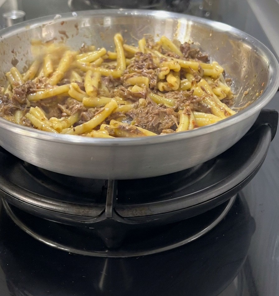 Casarecce-Pasta wird in einer Edelstahlpfanne unter eine dunkle, reichhaltige Sauce aus zerzupftem Fleisch und Zwiebeln gehoben.