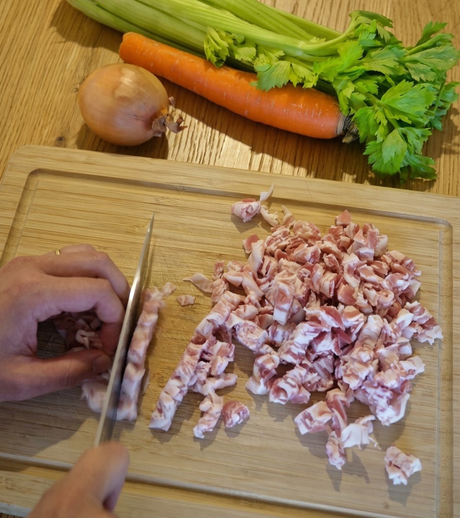 Nahaufnahme von Händen, die mit einem Kochmesser rosafarbene Pancetta-Streifen auf einem Holzbrett in kleine Würfel schneiden.