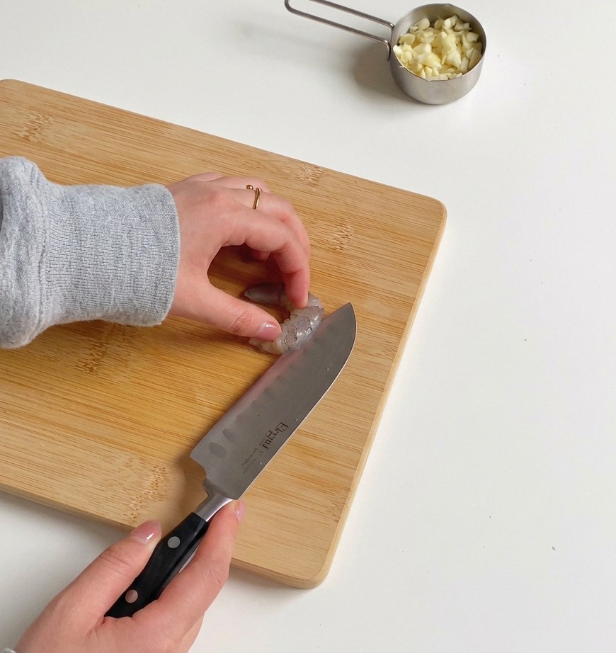 Nahaufnahme von Händen, die eine rohe, geschälte Garnele mit einem scharfen Messer auf einer Holzoberfläche im Schmetterlingsschnitt aufschneiden.