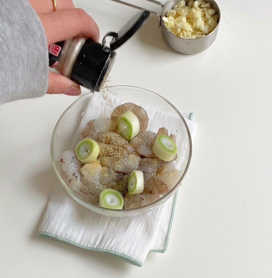 Eine Hand streut Salz und Pfeffer aus einem Gewürzstreuer über eine Schüssel mit rohen Garnelen und Frühlingszwiebelringen.