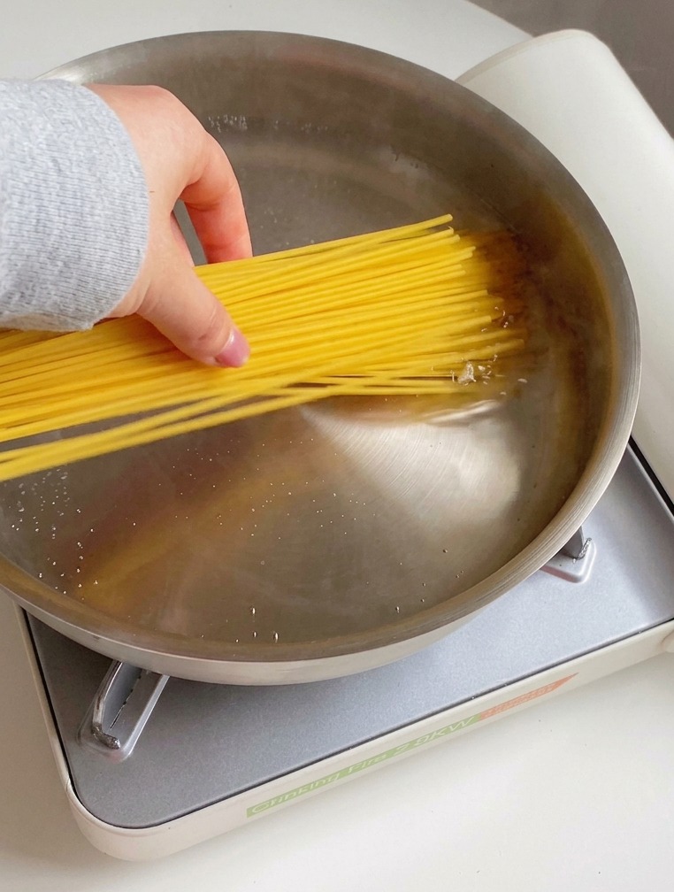 Eine Hand gibt ein Bündel trockene Spaghetti in eine Edelstahlpfanne mit kochendem Wasser, Öltropfen und Salz.