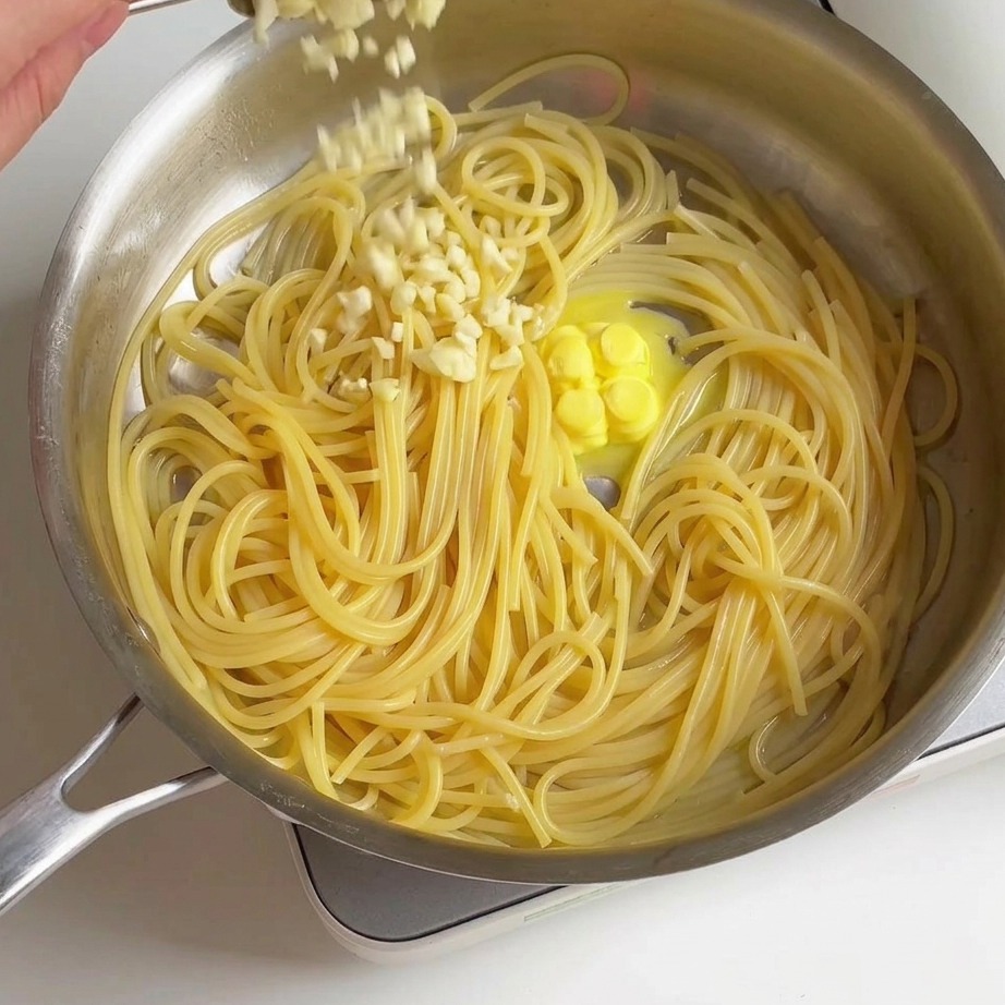 Frisch gehackter Knoblauch wird mit einem Löffel über gekochte Spaghetti und schmelzende Butter in einer Pfanne gestreut.