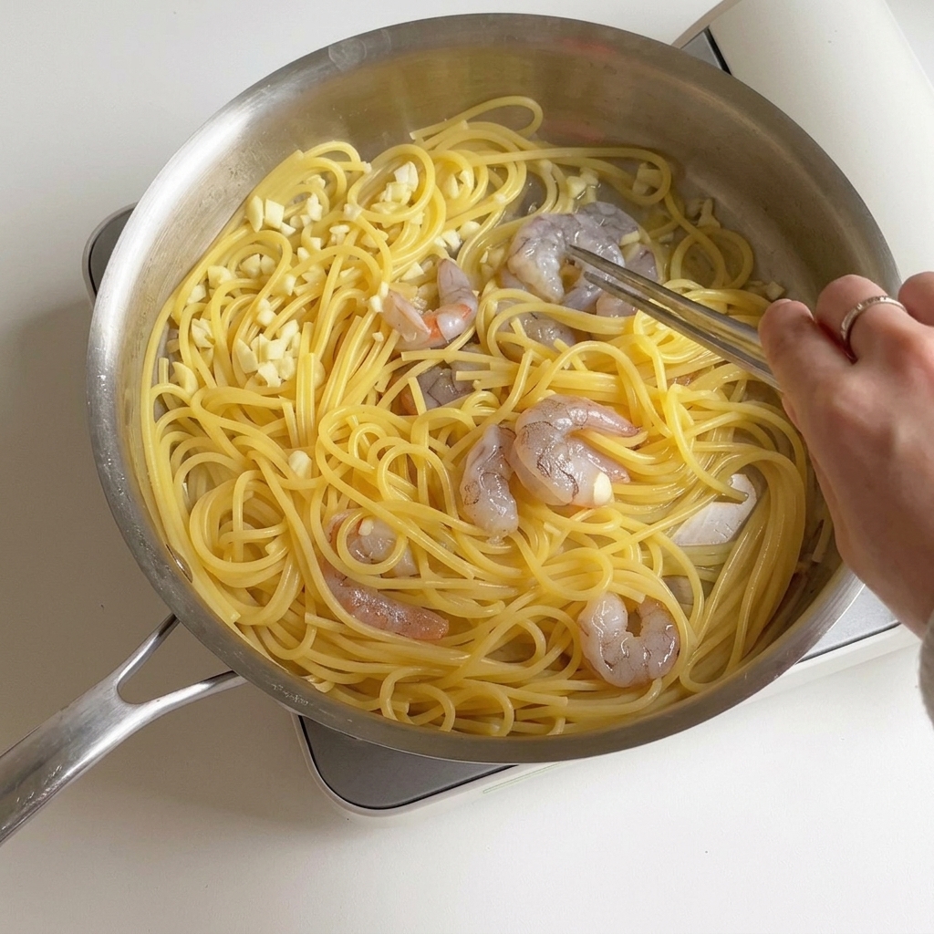 Eine Person brät Garnelen und Spaghetti in einer Pfanne an, während die Garnelen rosa und undurchsichtig werden.