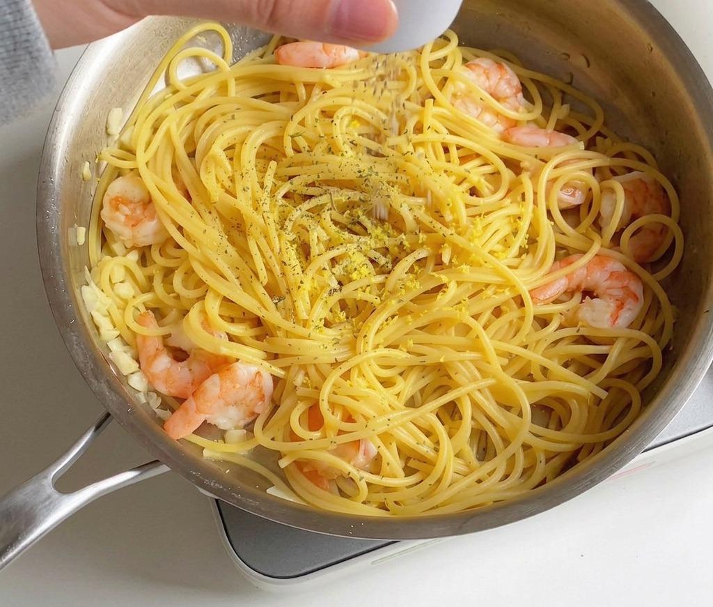 Eine Hand würzt eine Pfanne mit Garnelen-Zitronen-Spaghetti mit getrockneten Petersilienflocken und Salz.