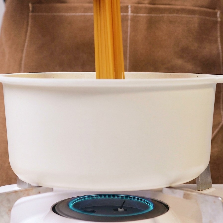 Eine Person gibt ein Bündel trockene Spaghetti in einen weißen Topf mit kochendem Wasser auf einem Gasherd.