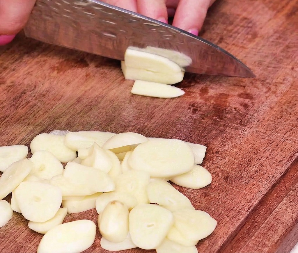 Nahaufnahme einer Hand, die Knoblauchzehen mit einem scharfen Kochmesser auf einem Holzbrett in gleichmäßige, dünne Scheiben schneidet.