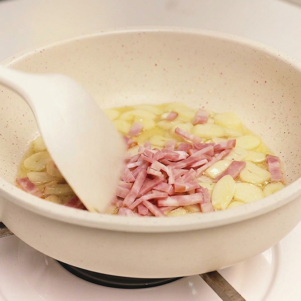 Ein weißer Pfannenwender rührt geschnittenen Speck und Knoblauchstücke in einer hellen Bratpfanne um.