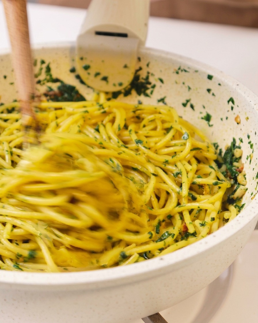 Spaghetti werden mit Holzstäbchen schwungvoll in einer Pfanne mit Kräutern und Öl vermengt.