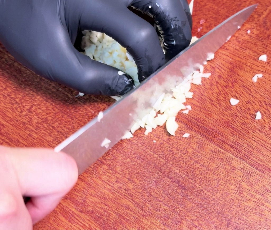 Eine Hand in einem schwarzen Handschuh hackt rohe Knoblauchzehen mit einem großen Kochmesser auf einem dunklen Holzschneidebrett fein.