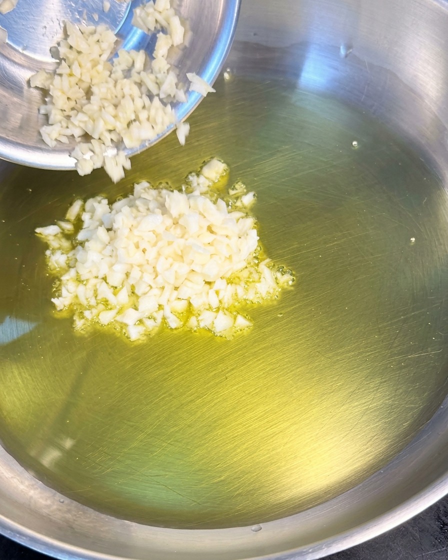 Gehackter weißer Knoblauch wird aus einer kleinen Metallschale in eine Pfanne mit heißem Olivenöl gegeben.