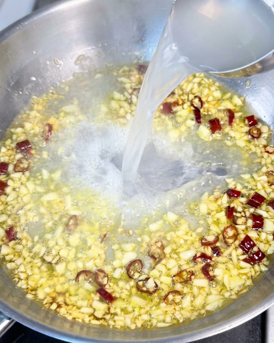 Trübes Nudelwasser wird mit einer Kelle in eine Pfanne mit Öl, Knoblauch und Chiliflocken gegossen.