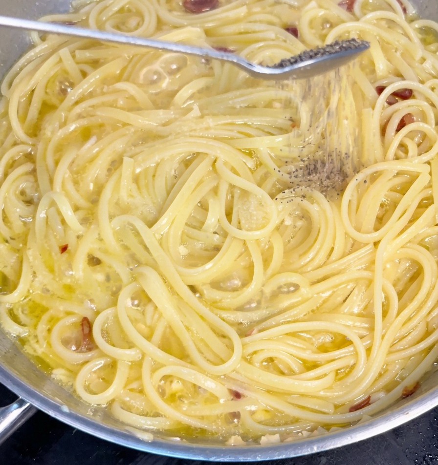 Frisch gemahlener schwarzer Pfeffer fällt von einem Löffel auf Spaghetti, die in einer Knoblauch-Öl-Sauce köcheln.