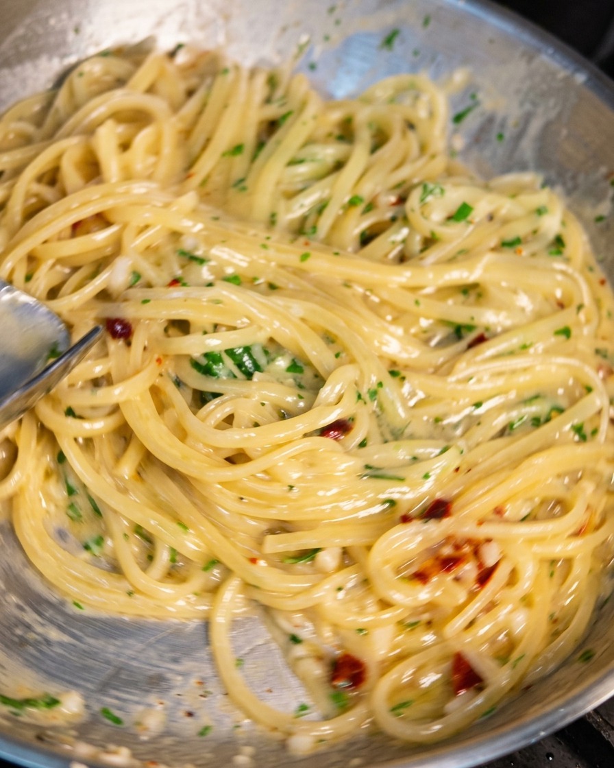 Eine Edelstahlpfanne voller Spaghetti wird mit einer Küchenzange geschwenkt. Die Nudeln are von einer glänzenden, cremigen Sauce überzogen, in der Knoblauchstücke, Chiliflocken und Petersilie zu sehen sind.