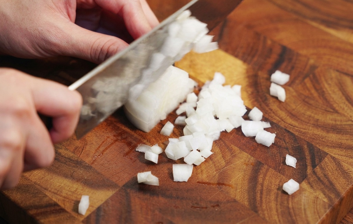 Hände schneiden eine weiße Zwiebel mit einem Kochmesser in feine Würfel auf einem Holzschneidebrett.
