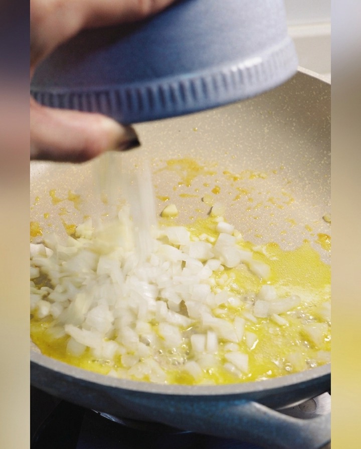 Gewürfelte weiße Zwiebeln werden aus einer blauen Schüssel in eine Pfanne mit geschmolzener Butter gegeben.