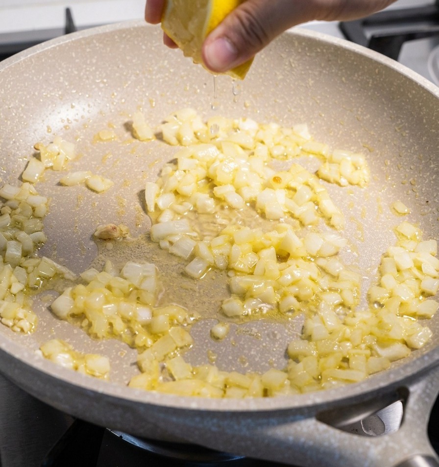 Eine Hand presst eine gelbe Zitronenhälfte über einer Pfanne mit glasig gedünsteten Zwiebel- und Knoblauchstücken aus.