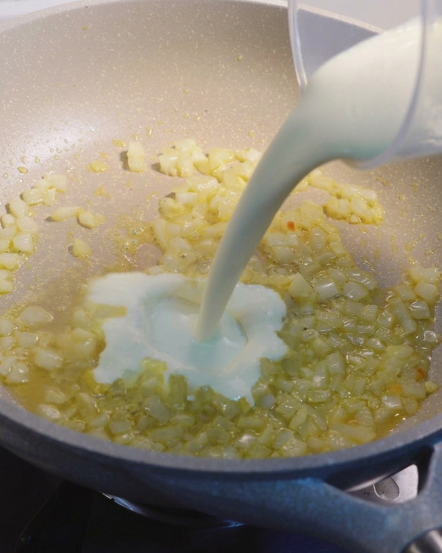 Flüssige Sahne wird aus einem Messbecher in eine gesprenkelte beige Pfanne mit gedünsteten Zwiebeln und Knoblauch gegossen.