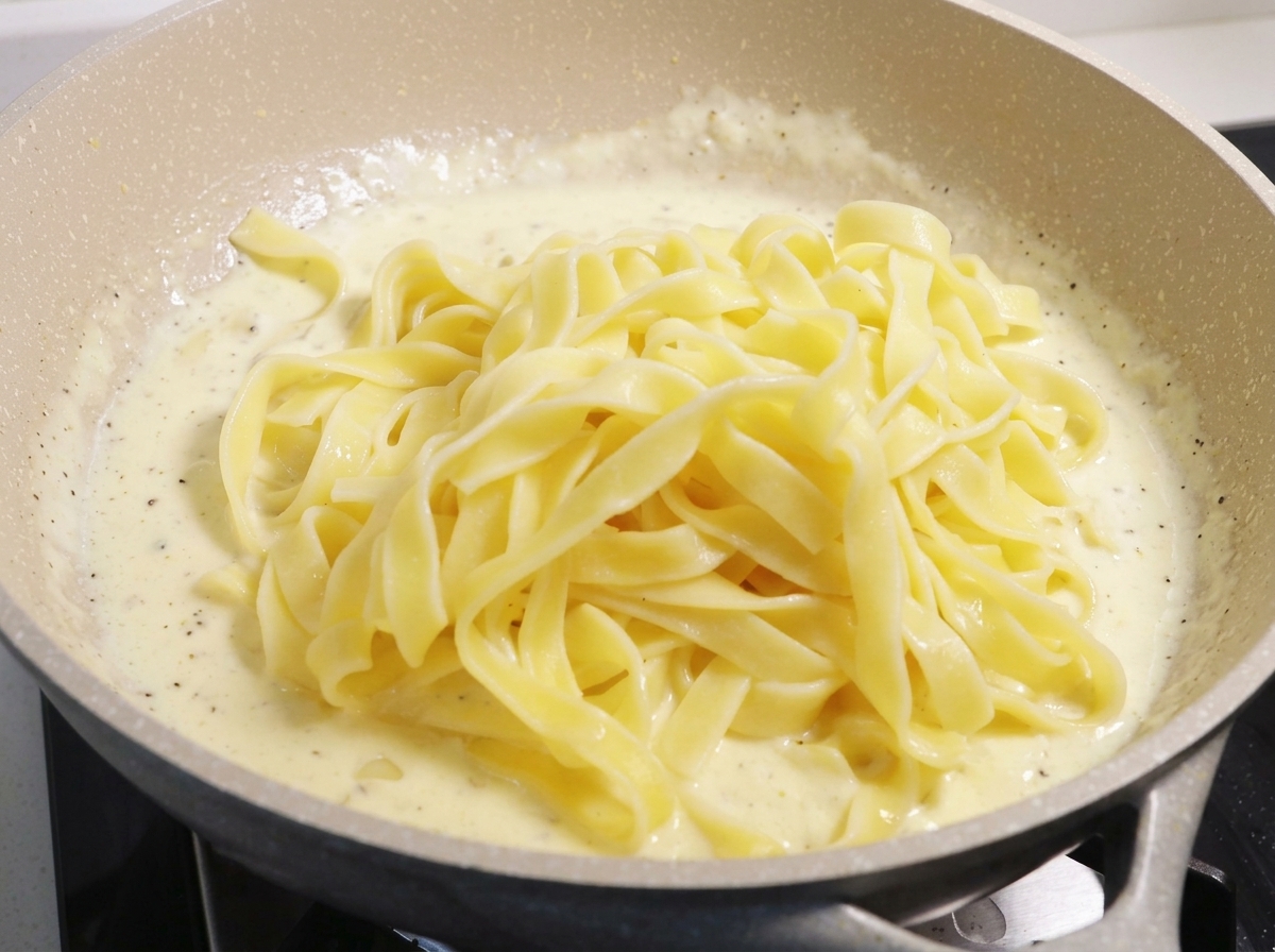 Ein großer Berg gekochter gelber Fettuccine-Nudeln liegt auf einer dickflüssigen, weiß brodelnden Sahnesauce in einer großen Pfanne.